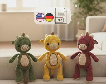 Op de afbeelding: Drie handgemaakte gehaakte koeienpoppen in groen, geel en rood. Elke pop heeft een witte buik, bruine hoorns en een lachend gezicht. De poppen zitten op een grijze bank.