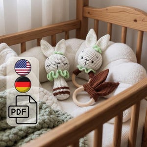 Puede incluir: Dos sonajeros de conejito de ganchillo hechos a mano en una cuna. Los conejitos son blancos con detalles marrones y verdes, y anillos de madera. La cuna es de madera clara y la ropa de cama es blanca y crema.