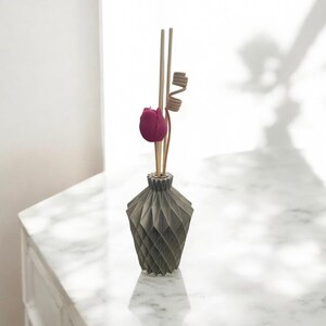 K&ouml;nnte beinhalten: Eine graue Diffusorflasche mit geometrischem Muster, einer dunkelrosa Tulpe und Holzst&auml;bchen. Der Diffusor steht auf einer wei&szlig;en Marmoroberfl&auml;che, mit einem Schatten im Hintergrund.