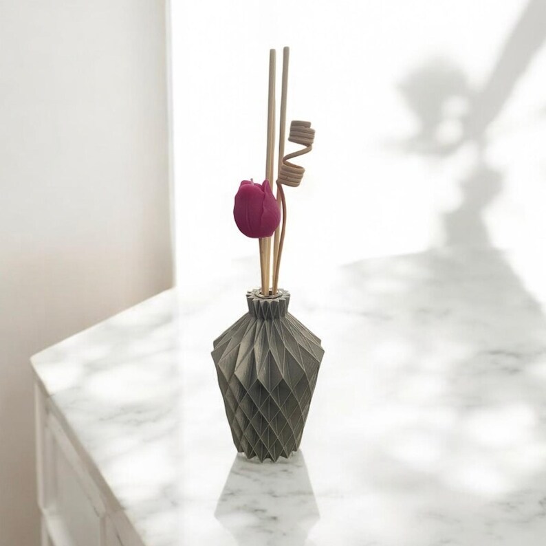 K&ouml;nnte beinhalten: Eine graue Diffusorflasche mit geometrischem Muster, einer dunkelrosa Tulpe und Holzst&auml;bchen. Der Diffusor steht auf einer wei&szlig;en Marmoroberfl&auml;che, mit einem Schatten im Hintergrund.