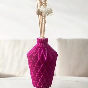 Puede incluir: Un jarrón geométrico magenta con un patrón de diamantes, que contiene palitos decorativos de madera, una flor blanca y una bola de madera. El jarrón está sobre una superficie reflectante.