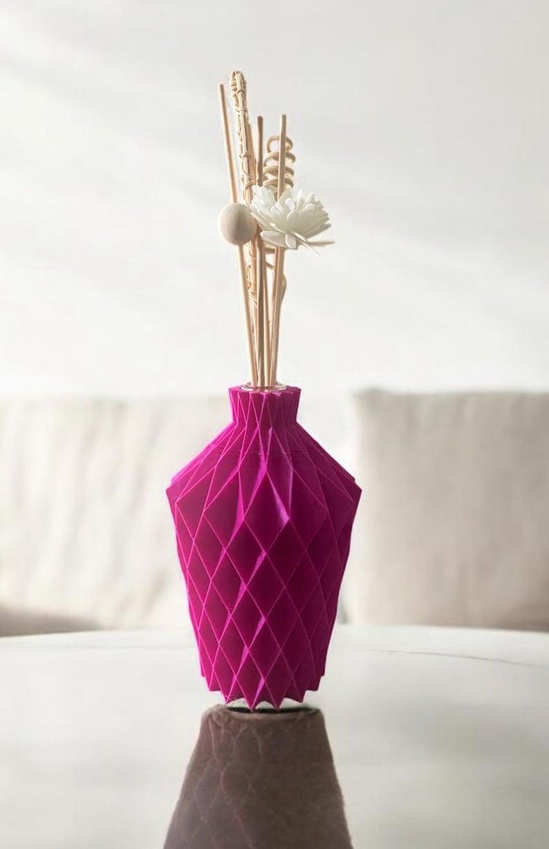 K&ouml;nnte beinhalten: Eine magenta geometrische Vase mit Rautenmuster, die dekorative Holzst&auml;be, eine wei&szlig;e Blume und eine Holzkugel enth&auml;lt. Die Vase steht auf einer reflektierenden Oberfl&auml;che.