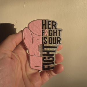 May include: A pink boxing glove design with the text "HER FIGHT IS OUR FIGHT" in black lettering. A pink ribbon is included in the text. The design has a glittery silver border.