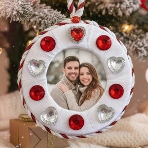 May include: A white and red Christmas ornament with a photo of a couple. The ornament is decorated with red and silver heart-shaped and round gems. It hangs from a red and white twisted cord. The background includes a Christmas tree and gifts.