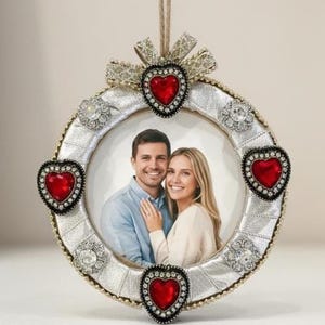 May include: A round silver photo frame with a couple's photo inside. The frame is decorated with red heart-shaped jewels and clear rhinestones. A gold bow and a twine loop are at the top for hanging.