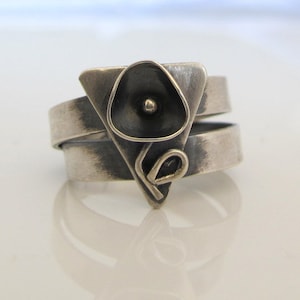 Op de afbeelding: Een zilveren ring met een geometrisch ontwerp met een zwart bloemachtig element. De ring is gemaakt van twee banden die in elkaar gedraaid zijn.