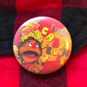 Op de afbeelding: Een ronde, rode button met de tekst "I've got CABIN FEVER" in gele en oranje letters. De button toont een cartoonfiguur met een oranje huid, een wijd open mond en een gele bloemenkrans.