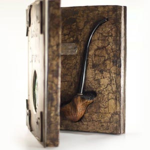 Op de afbeelding: Een houten pijp tentoongesteld in een decoratieve doos. De doos is bruin met een getextureerd oppervlak en een scharnierende deur. De pijp heeft een gebogen steel en een bruine ketel. De doos heeft metalen accenten en tekst.