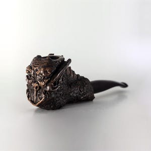 以下が含まれることがあります： 精巧に彫刻された、濃い茶色のパイプ。ボウルは、グロテスクな顔の、詳細で質感のあるデザインが特徴です。ステムは滑らかで湾曲した濃い茶色です。パイプは無地の白い背景に置かれています。