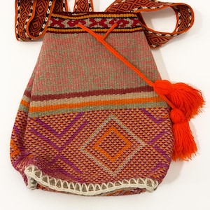 May include: A woven shoulder bag with a geometric pattern in shades of orange, purple, and pink. The bag has a matching strap and a bright orange tassel. The bag's design features a diamond pattern.