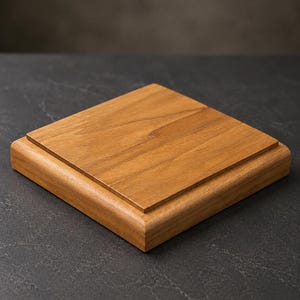 May include: A square wooden display base with a raised top. The base is made of light brown wood and has a smooth, polished finish. The edges are rounded, and it sits on a dark surface.