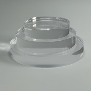 May include: A clear acrylic display stand featuring three circular levels. The stand is designed for product presentation, with each tier reducing in size upwards. The transparent material provides a clean, contemporary look.