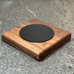 Può includere: Un sottobicchiere quadrato in legno con finitura marrone scuro e inserto circolare nero. Il sottobicchiere è realizzato in legno massello e ha una superficie liscia e lucida. L'inserto nero è centrato sul sottobicchiere.