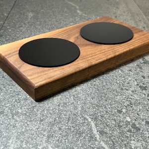 Può includere: Un set di sottobicchieri in legno con una base rettangolare in noce e due sottobicchieri neri circolari. La venatura del legno è visibile. Il set è progettato per due bevande.