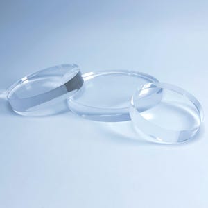Puede incluir: Tres discos circulares transparentes de acrílico de diferentes tamaños están dispuestos sobre una superficie azul claro. Los discos son transparentes, permitiendo el paso de la luz, y tienen un aspecto liso y pulido. La disposición sugiere un elemento de exhibición o decorativo.