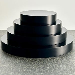 May include: A tiered, black, circular display stand. The stand is composed of four stacked, solid, black discs of varying diameters, creating a stepped effect. The surfaces are smooth and reflect light, giving a polished appearance. The stand is suitable for displaying items.