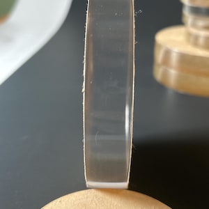 May include: A transparent, circular acrylic disc is shown on its edge, revealing its thickness. It rests on a light brown, circular base. The background is dark grey, with a hint of another similar object.