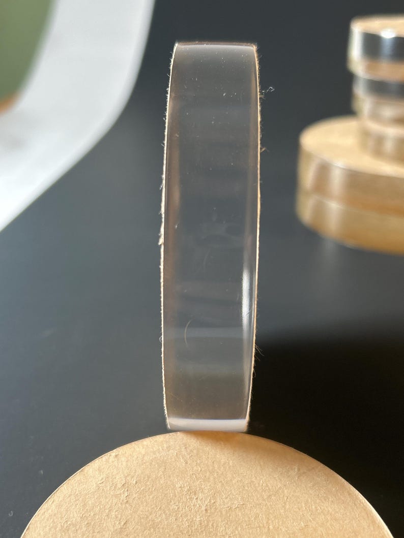 May include: A transparent, circular acrylic disc is shown on its edge, revealing its thickness. It rests on a light brown, circular base. The background is dark grey, with a hint of another similar object.