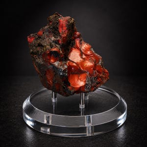 May include: A raw, irregular mineral specimen featuring vibrant orange-red and deep black coloration with a glossy, glass-like texture. The stone is mounted on a clear, circular acrylic display stand supported by four small, silver-toned metal pillars.