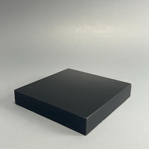 May include: A solid black square block with a smooth, matte finish. The block is a uniform colour and shape, with clean, straight edges. The object is likely a display stand or base.