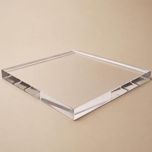 May include: A clear, square acrylic block with beveled edges. The transparent material reflects light, creating a subtle play of shadows. The block is designed with clean lines and a smooth surface.