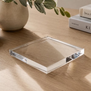 May include: A clear, square acrylic block with a beveled edge sits on a light wood surface. The block reflects light, creating a prism effect. A decorative vase, a wooden bowl, and books are in the background.