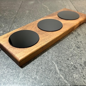Può includere: Un vassoio di legno con tre sottobicchieri neri circolari. Il vassoio è realizzato in legno marrone chiaro con venature visibili. I sottobicchieri sono uniformemente distanziati sulla superficie del vassoio.