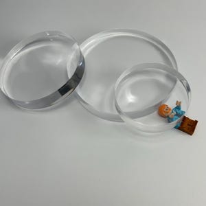 May include: Transparent acrylic circles in different sizes. A small toy figure is inside one of the circles. The circles are clear and appear to be made of a thick material. The objects are on a white surface.