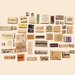 May include: A collection of wooden stamps with various inspirational quotes and designs. The stamps feature phrases like "Laughter," "Believe," and "Wings and Fly." Some stamps have floral and peace sign motifs. The stamps are in various sizes and shapes.