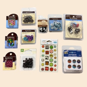 May include: A collection of Recollections brand craft supplies, including brads, eyelets, and decorative stickers. The items are packaged in clear plastic containers and feature various colors and designs, perfect for scrapbooking and paper crafts.