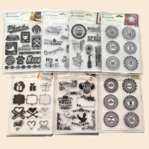 May include: A collection of clear stamps in plastic packaging. The stamps feature various designs, including text, circles, flowers, butterflies, and decorative elements. Some stamps have text like "Meow", "Happy Birthday", and "Beautiful".