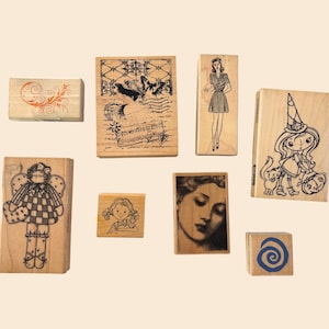 May include: A collection of wooden stamps with various designs. The stamps feature images of a butterfly, a woman in a dress, a girl, a witch, and an angel. One stamp has an orange floral design, and another has a blue spiral.