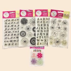 May include: Clear stamps in various designs, including letters, flowers, and decorative elements. The stamps are packaged in clear plastic with colorful labels. The text "SCRAPPY CAT" and "Clear Stamps" are visible on the packaging.