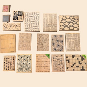 May include: A collection of wooden stamps with various designs, including floral patterns, geometric shapes, newspaper print, and a crossword puzzle. The stamps are in various sizes and shapes, with a light brown wooden base.