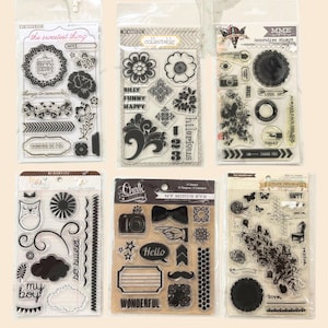 May include: Six clear acrylic stamp sets with black designs. The sets feature floral, decorative, and word elements like "Happy," "Hello," and "Wonderful." The stamps are packaged in clear plastic with cardboard headers, ideal for scrapbooking and crafting.