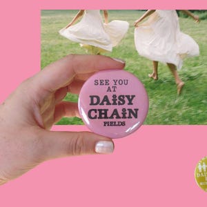Puede incluir: Un botón redondo de color rosa con el texto negro SEE YOU AT DAISY CHAIN FIELDS, donde pequeños iconos de flores reemplazan la letra I en DAISY y CHAIN. El botón se muestra sobre un fondo que muestra a dos personas con vestidos blancos.