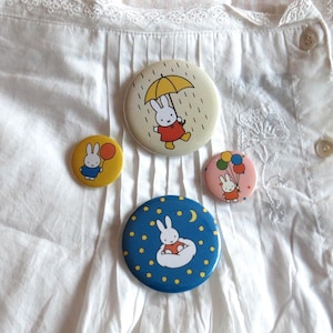 May include: Four circular pinback buttons featuring the Miffy character. One large cream button shows Miffy with a yellow umbrella in rain. A large blue button shows Miffy on a cloud with stars. Two smaller buttons show Miffy with balloons.