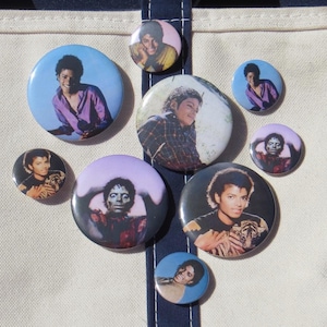May include: A collection of eight circular pin-back buttons featuring various photographic portraits of Michael Jackson. The buttons vary in size and display the artist in different outfits, including a purple shirt, a plaid shirt, and a tiger print.