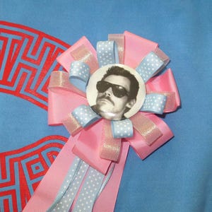 May include: A circular pinback button featuring a black and white portrait of a person wearing sunglasses, centered on a rosette made of layered pink grosgrain ribbon and light blue ribbon with white polka dots, with three long ribbon tails.