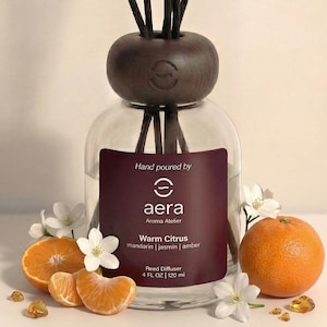 May include: A clear glass reed diffuser with a dark brown wooden top and reeds. The label reads "Warm Citrus" and "4 FL OZ | 120 ml." The image includes orange slices, a whole orange, and white flowers, suggesting a citrus fragrance.