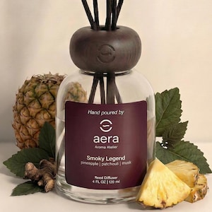 May include: A clear glass reed diffuser with a dark brown wooden top and black reeds. The label reads "Smoky Legend" with pineapple, patchouli, and musk scents. The diffuser is surrounded by pineapple slices, a whole pineapple, and green leaves.