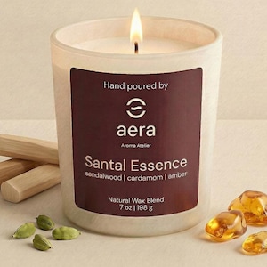 May include: A lit candle in a frosted glass jar with a dark maroon label reading Hand poured by aera Aroma Atelier Santal Essence sandalwood | cardamom | amber Natural Wax Blend 7 oz | 198 g, surrounded by wooden sticks, green cardamom pods, and amber stones.