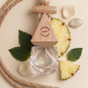 May include: A clear glass perfume bottle with a wooden top, a triangle-shaped wooden piece with a logo, and a decorative rope. The image includes pineapple slices, green leaves, and small white stones, all arranged on a light beige surface.