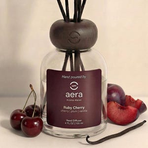 May include: A clear glass reed diffuser with a dark brown wooden top and reeds. The label reads "Ruby Cherry" with cherry, plum, and vanilla scents. Fresh cherries, plum slices, and a vanilla bean are arranged around the diffuser. The bottle contains 4 fl oz or 120 ml.