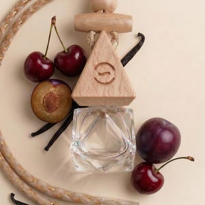 May include: A clear glass perfume bottle with a wooden triangle top, surrounded by fresh cherries, plums, and vanilla beans. The wooden top has a carved design. A braided rope with gold accents is also present.