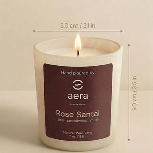 May include: A lit candle in a frosted glass jar with a dark label reading Hand poured by aera Aroma Atelier Rose Santal rose | sandalwood | musk Natural Wax Blend 7 oz. The jar measures 3.1 inches wide at the top, 2.9 inches at the base, and 3.5 inches tall.
