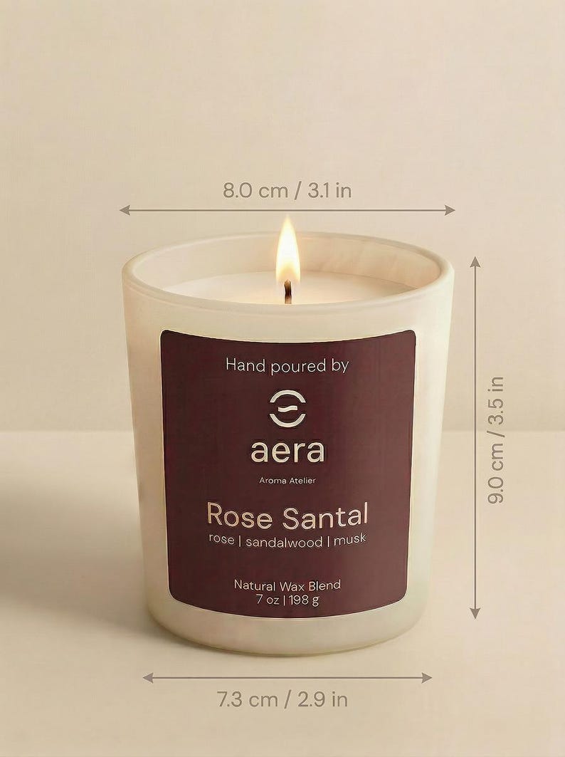 May include: A lit candle in a frosted glass jar with a dark label reading Hand poured by aera Aroma Atelier Rose Santal rose | sandalwood | musk Natural Wax Blend 7 oz. The jar measures 3.1 inches wide at the top, 2.9 inches at the base, and 3.5 inches tall.