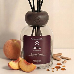 Może przedstawiać: Szklany dyfuzor trzcinowy z ciemnobrązową drewnianą górą i trzcinami. Etykieta głosi "Cognac Peach" i "120 ml". Brzoskwinia i plasterki brzoskwini leżą obok dyfuzora. Dyfuzor jest z "aera Aroma Atelier."
