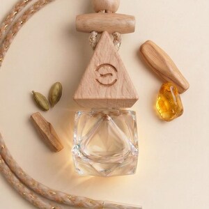 May include: An air freshener with a clear glass container and a wooden triangle top with a carved design. The air freshener is suspended by a beige and gold rope. Additional wooden pieces and amber-colored stone are arranged around it.