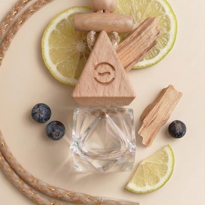 May include: A clear, geometric glass bottle with a wooden triangle top, a braided rope, and a wooden accent. The image includes lemon slices, juniper berries, and wood pieces. The wooden triangle has a circular logo. The overall theme is aromatherapy.